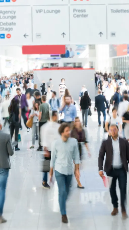 A busy, brightly lit convention center hall with a crowd of people in motion and overhead signs for a VIP Lounge and Press Center.